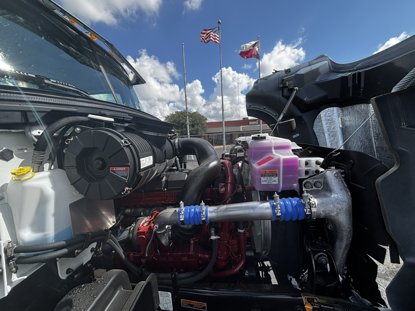 Engine bay of new 2027 Mack GR64FT daycab showing turbocharger, coolant reservoir, pink coolant/DEF reservoir, hoses, radiator fan shroud, and engine block.