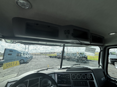 Interior cab view of a 2027 Mack GR64FT from driver’s seat showing dashboard gauges, steering wheel, overhead module, and windshield with other trucks outside.