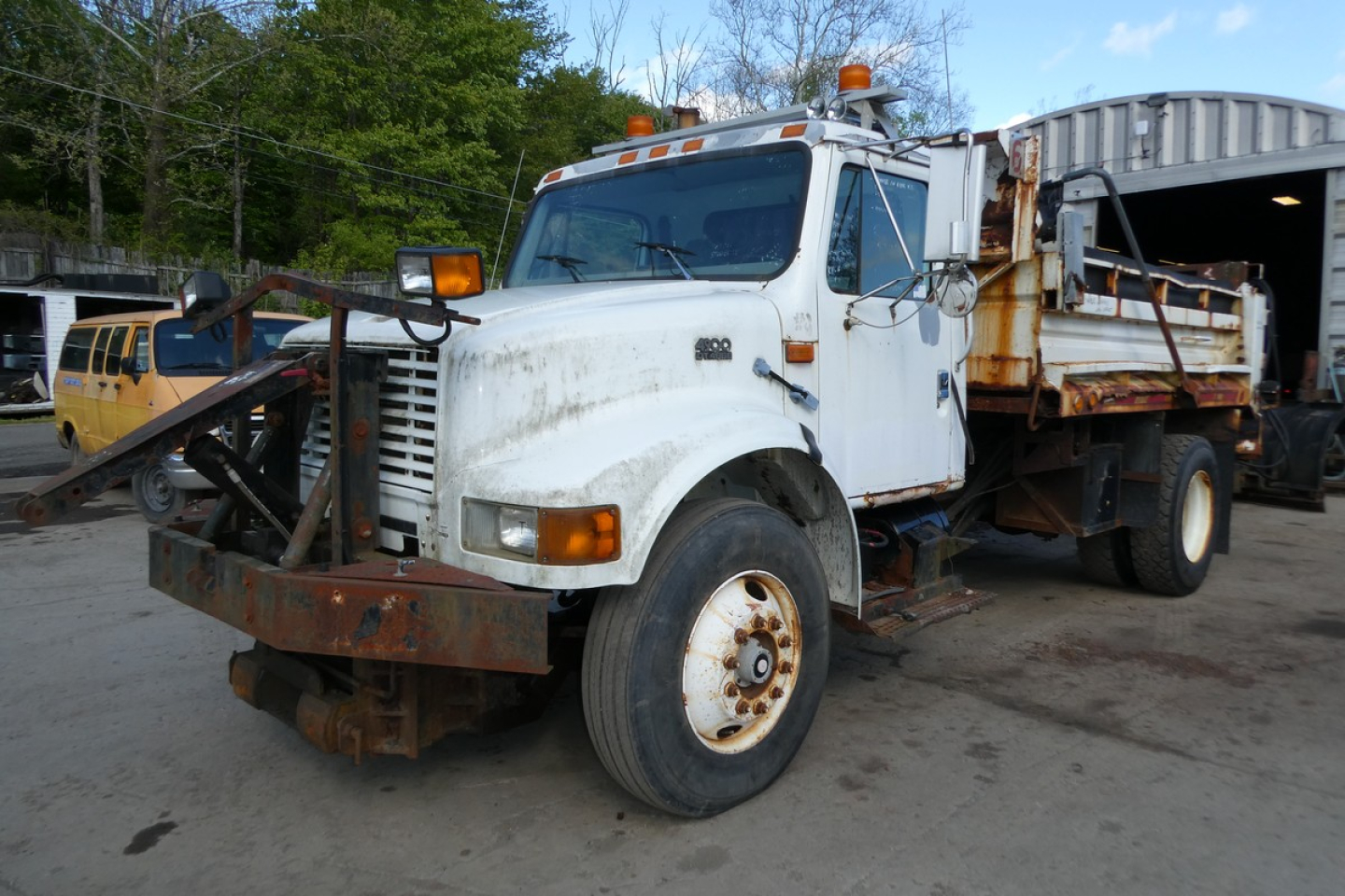 Dump truck front-left view of a white 1998 International 4900 with a rusted front bumper, orange warning beacon, and dirt/grime on body.