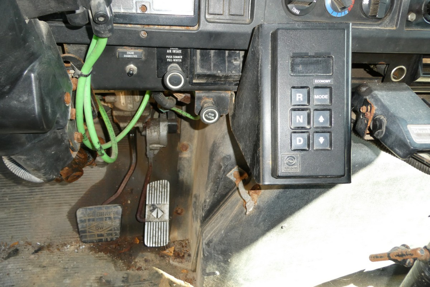 Dashboard area of a truck showing pedals, green wiring, and a shifter panel with R N D P buttons. Visible labels include gear selector block and some switches.