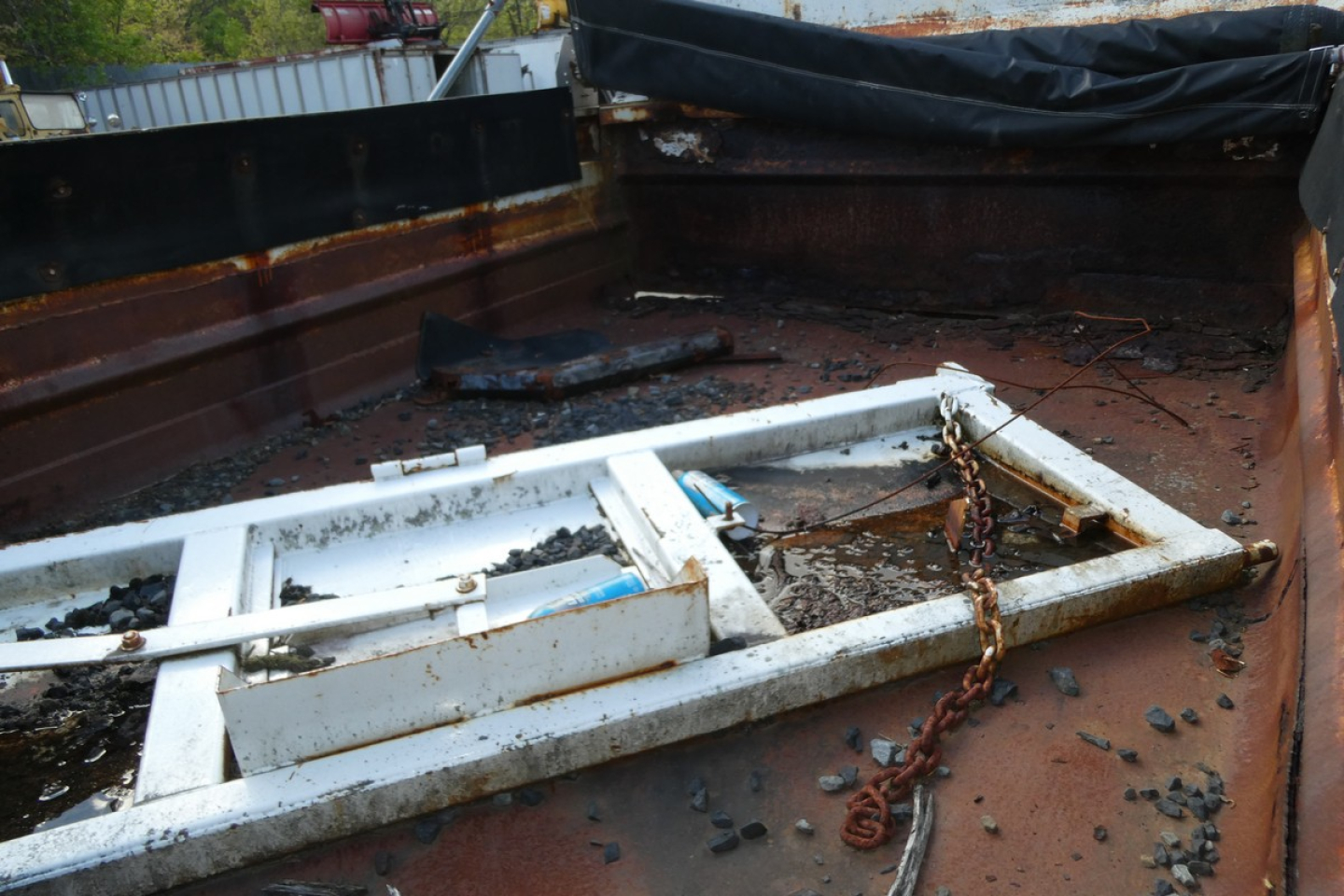 Dump truck bed with a metal frame or equipment frame lying inside, chained and resting on a rusted surface inside the truck bed.