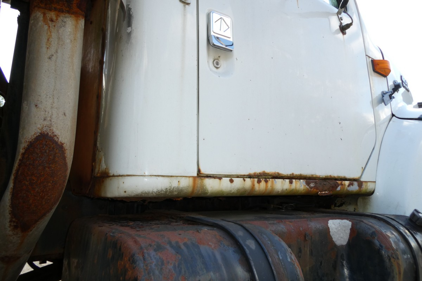 Truck cab with visible door, emblem, and rust on lower cab edge; partial view of fuel tank and exhaust area.