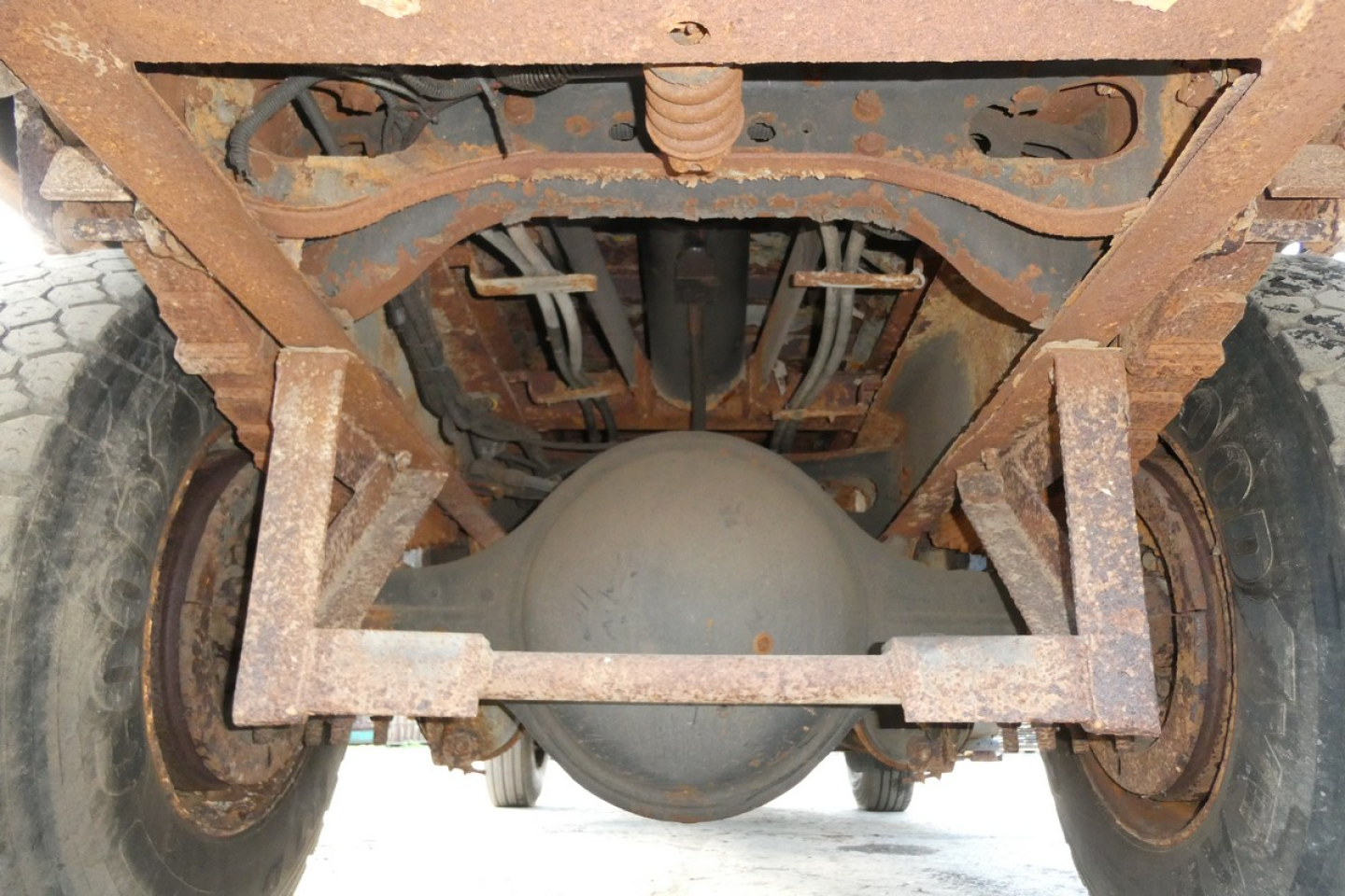 Underbody view of a dump truck showing rear axle housing, leaf springs, and frame rails with rust; visible components include differential housing and suspension. No legible brand text identified.