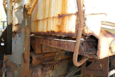 Heavy rusted metal frame and damaged components on a dump truck chassis, with corroded beams and hydraulic lines visible.
