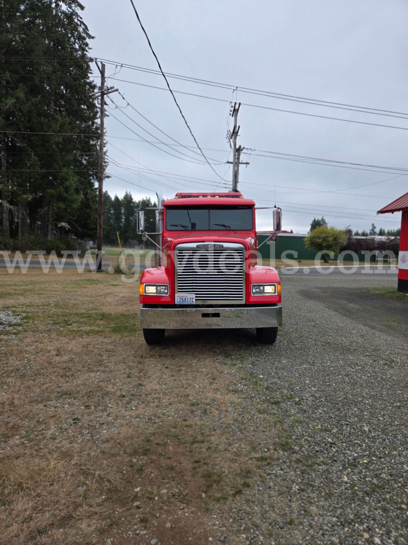 Front view of a red Freightliner FLD120 truck on a gravel lot, headlights, grille, bumper visible with a flat surroundings.
