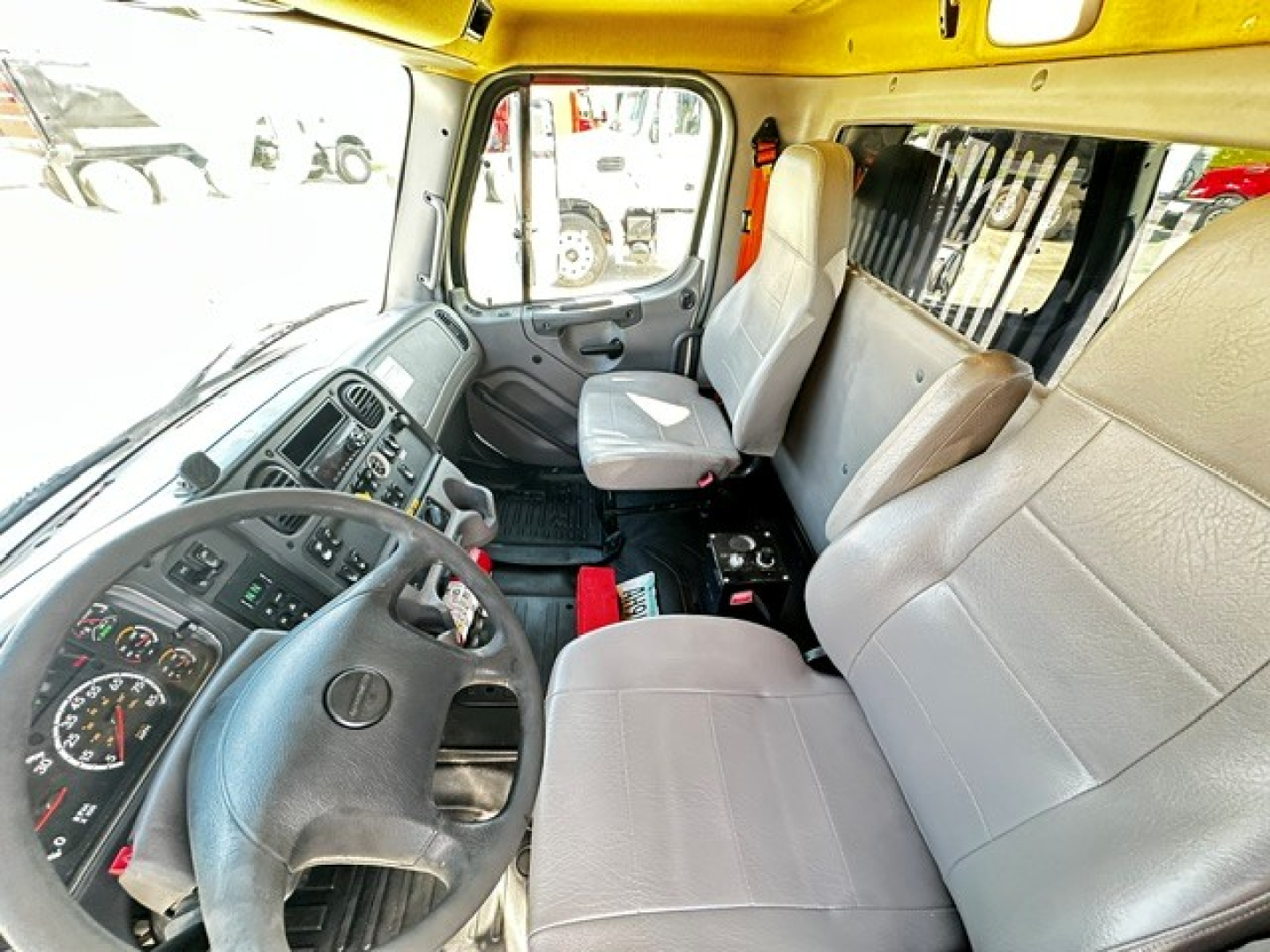 Cab interior of a 2018 Freightliner M2106 rollback truck showing steering wheel, dashboard, driver seat, and passenger seat in beige/gray fabric with yellow exterior frame.