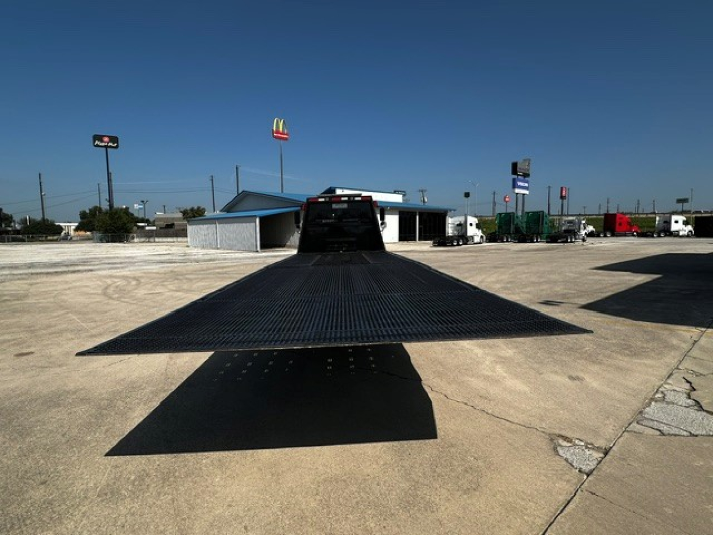 Tow truck flatbed ramp extended in a dealership lot, Freightliner M2106 identification and surrounding trucks in distance.