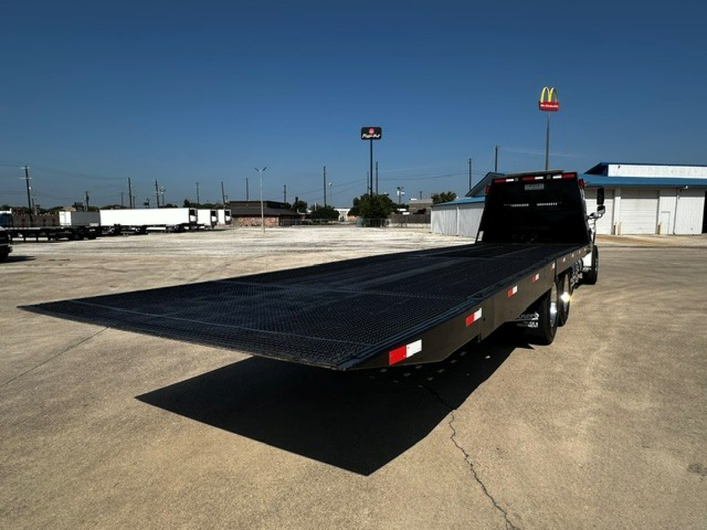 Flatbed tow truck with extended platform in a parking lot, Freightliner M2106 body style, visible cab at rear, long black steel bed, no visible text other than generic markings.