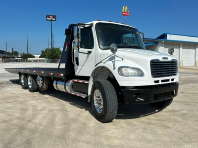 Flatbed tow truck with white cab and black bumper, Freightliner M2106, side view showing dual rear axles, flatbed, and towing equipment.