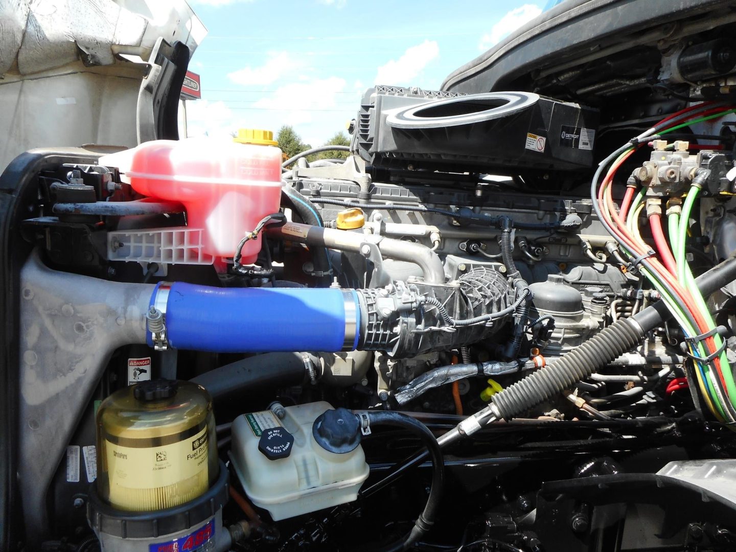 Freightliner Cascadia engine bay showing coolant reservoir, power steering cooler, and numerous hoses and wiring harnesses.