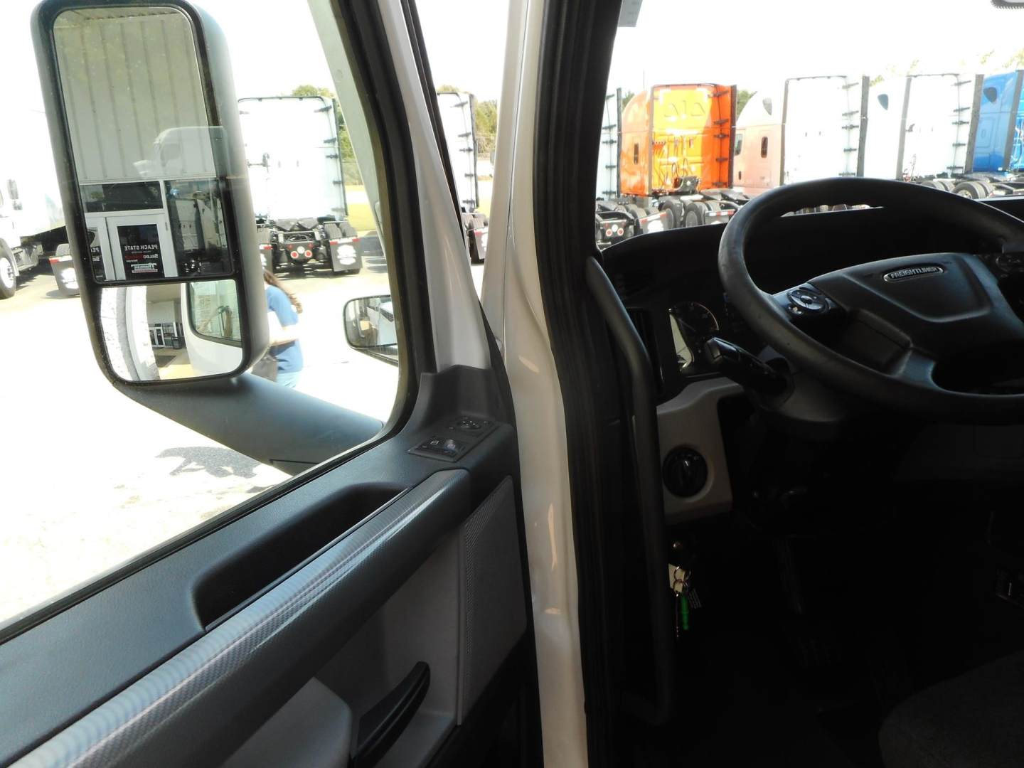 Freightliner Cascadia interior view from driver door showing steering wheel and dashboard; side mirror and window frame; exterior trucks in lot visible.