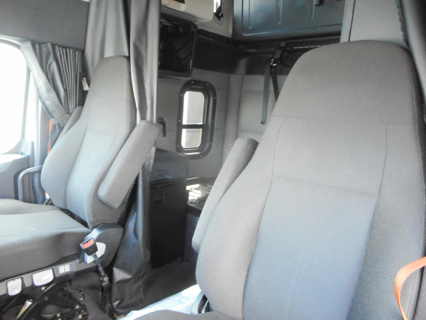Cab interior of a Freightliner Cascadia truck showing two gray seats and a doorway with window; dashboard and overhead storage visible.