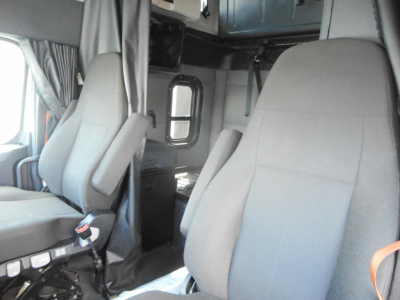 Cab interior of a Freightliner Cascadia truck showing two gray seats and a doorway with window; dashboard and overhead storage visible.