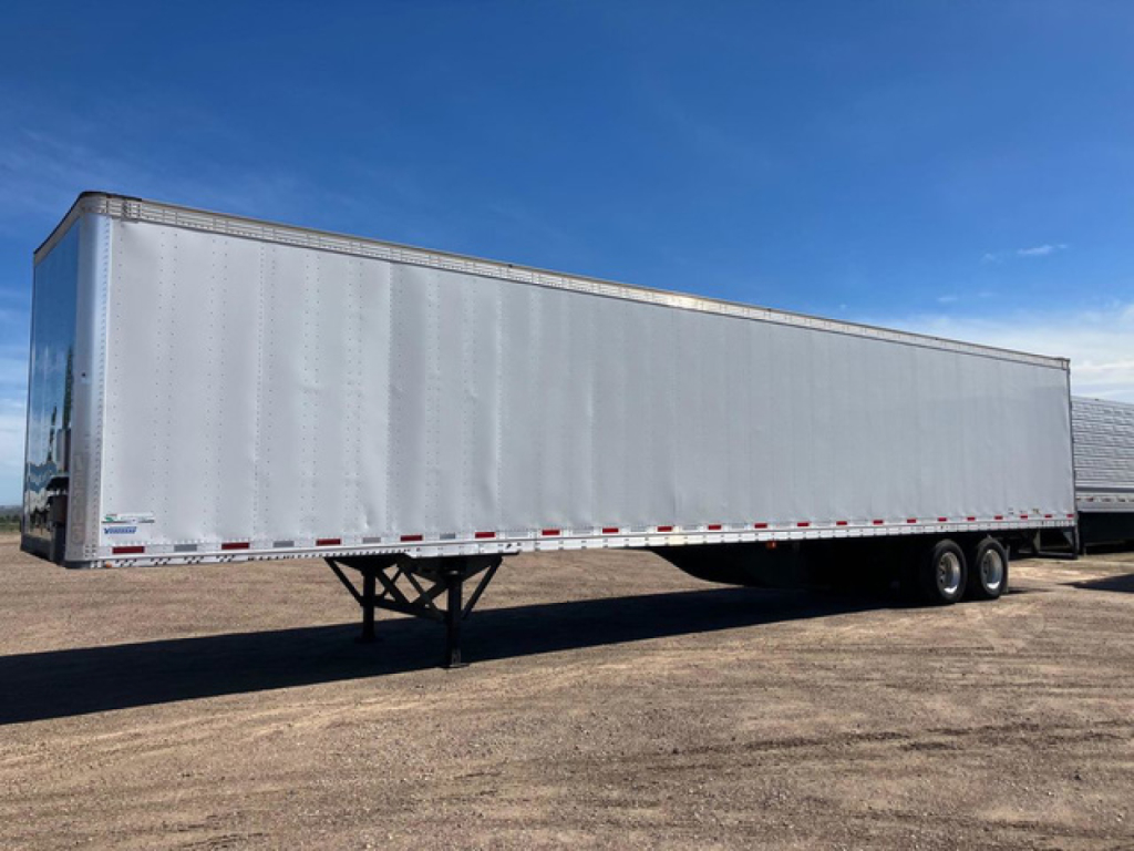 Semi trailer in side view showing a white dry van trailer with dual rear wheels and reflective tape along the sides.