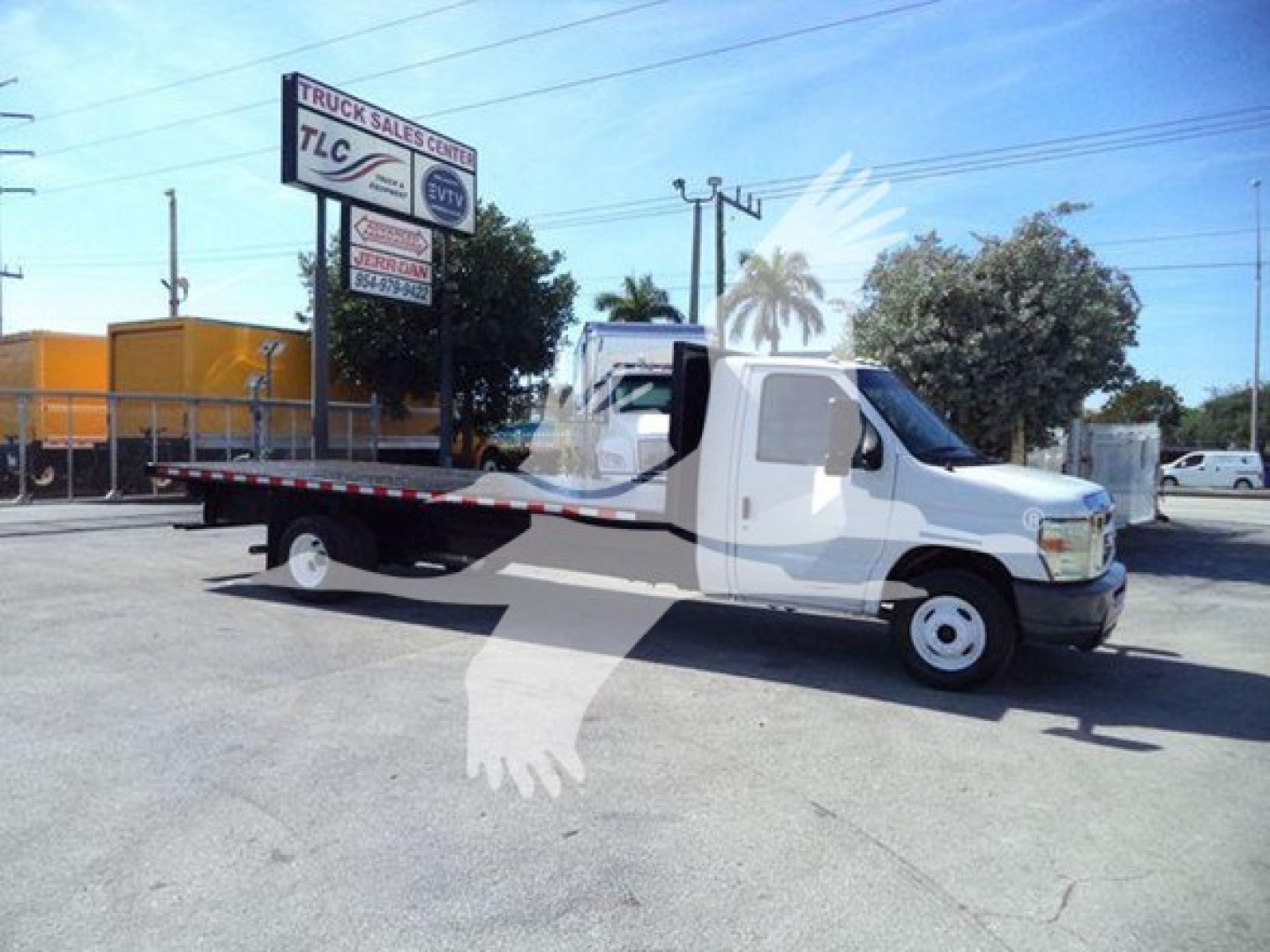Flatbed tow truck with white cab and long flatbed, Ford E-Series badge not visible, no visible text on side; background shows TLC truck sales center sign and palm tree watermark.