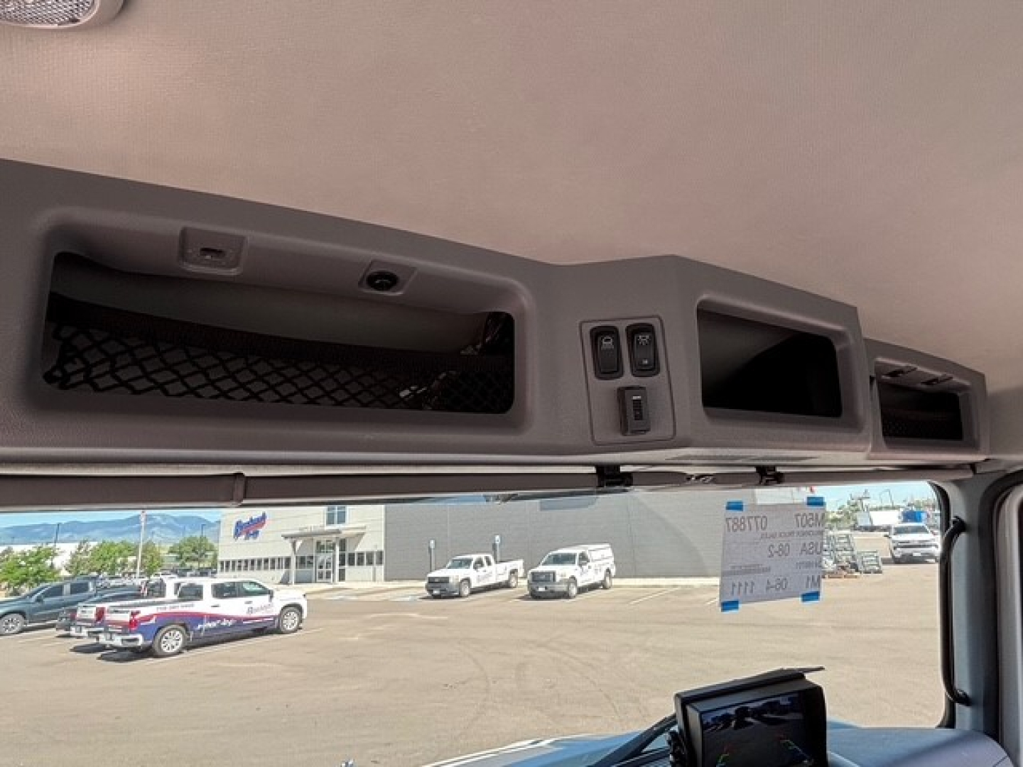 Vehicle cabin overhead panels and view through windshield of a new 2026 Mack MD7 truck showing interior dashboard area and exterior lot.