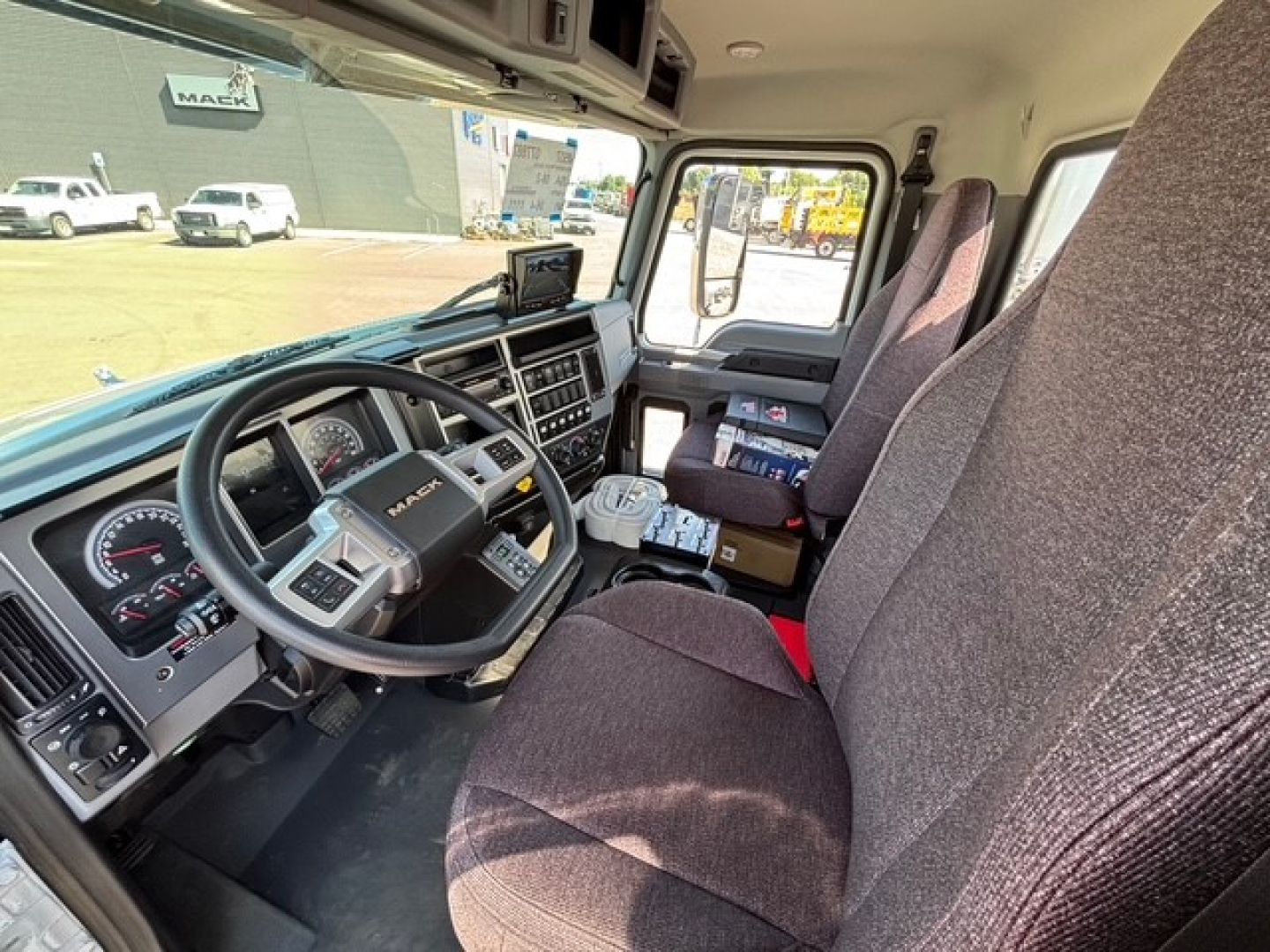 Interior view of a 2026 Mack MD7 truck cab showing steering wheel, dashboard gauges, and driver seat with fabric upholstery.