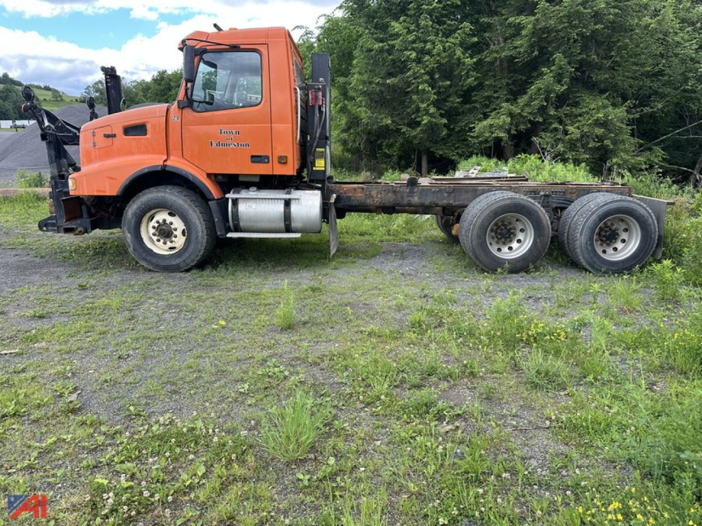 Orange Volvo VHD cab and chassis truck with flatbed rail and tandem rear axles, showing VIN/brand decals on door and engine bay; industrial lot with grass and trees.