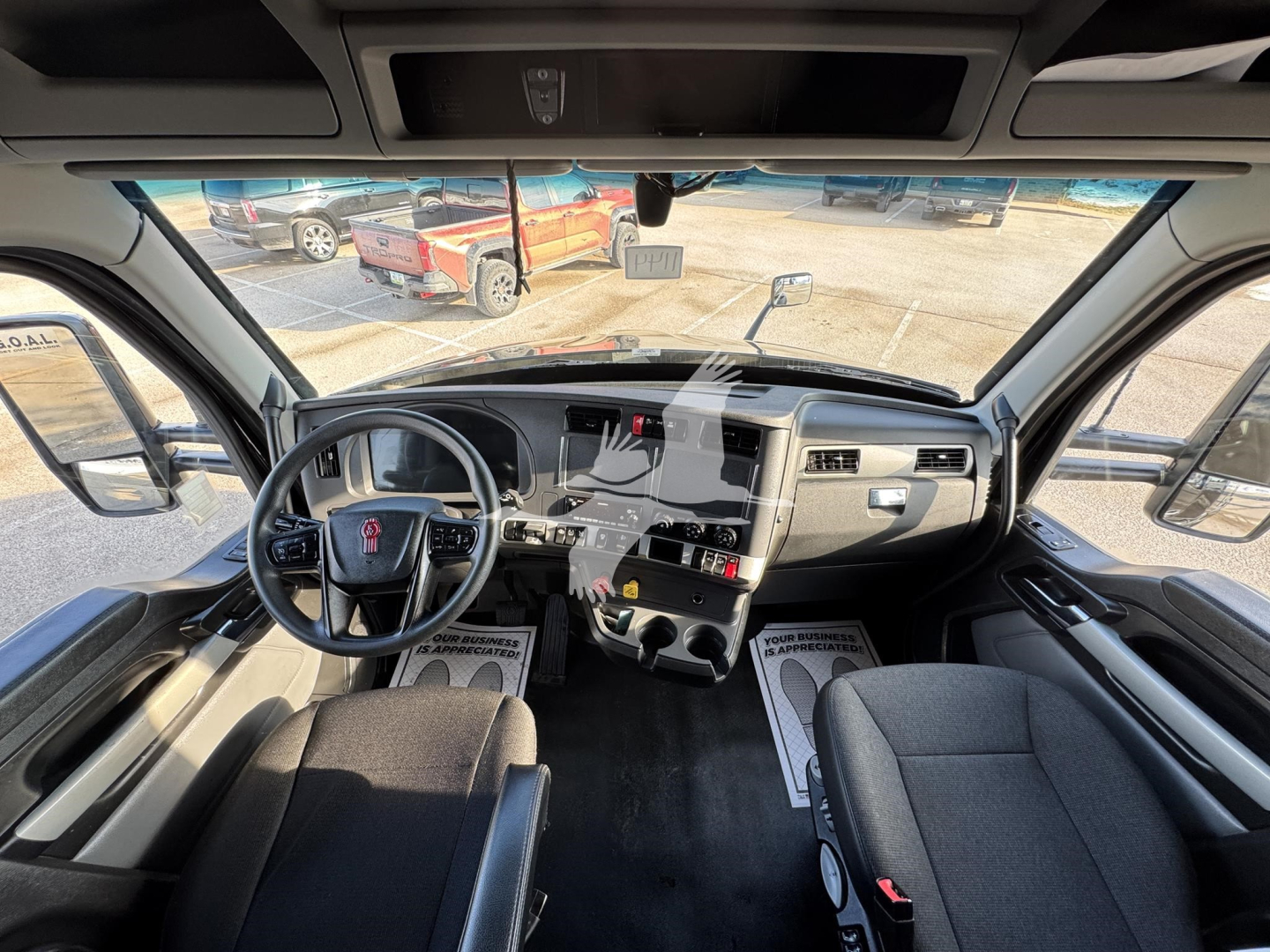 Truck cab interior showing steering wheel, dashboard with center console and two front seats; windshield view of yard with other trucks.