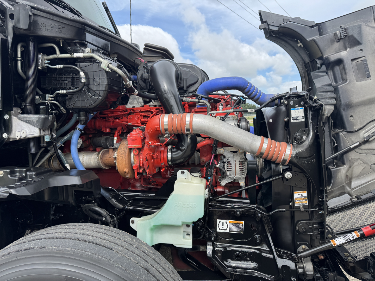 Kenworth T680 engine bay with red engine, blue hoses, silver intake, and black frame; visible coolant reservoir and various piping.