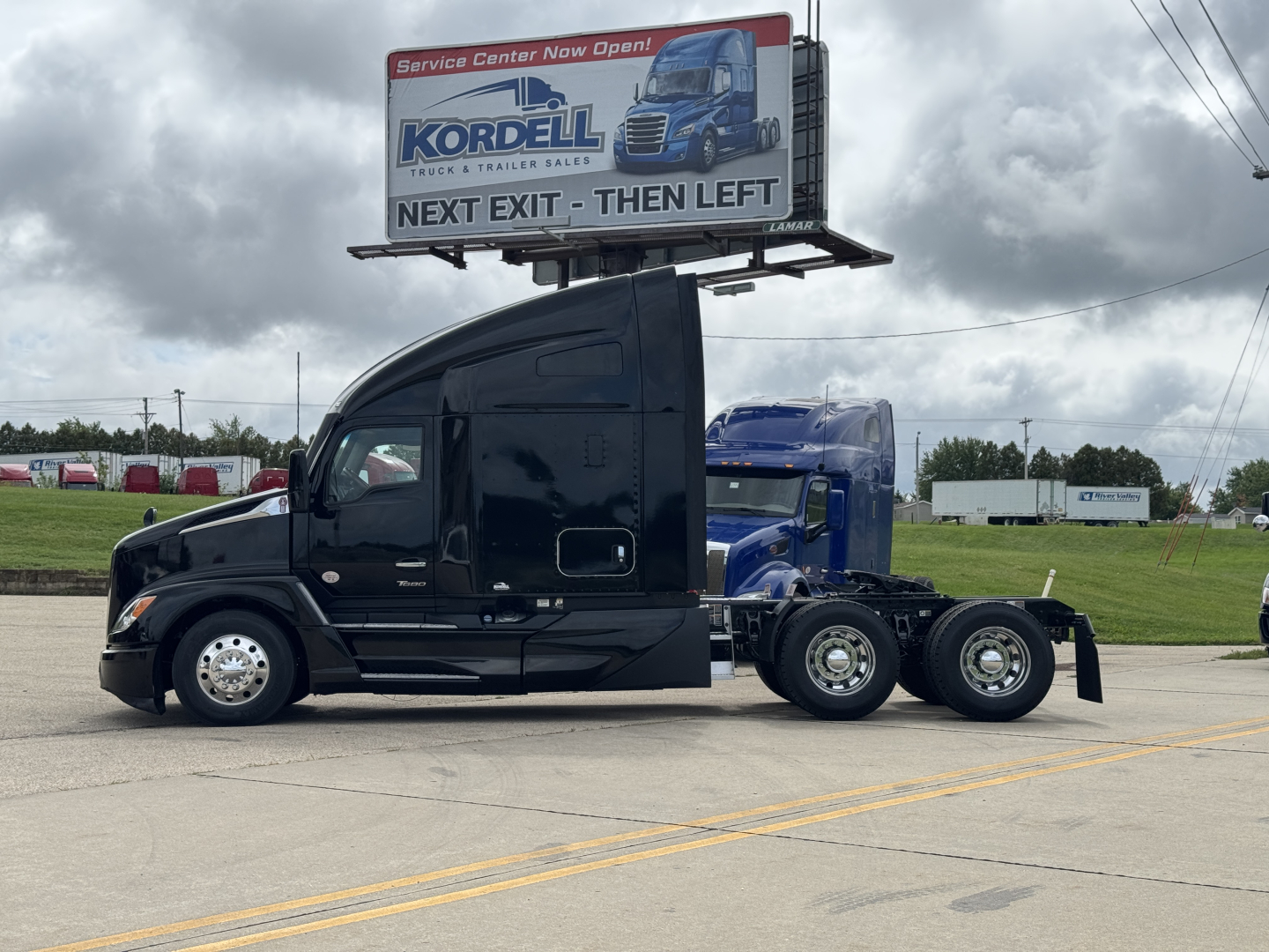 Kenworth T680 tractor in side profile view, black cab with sleeper, dual rear axles, billboard in background, dealership lot.