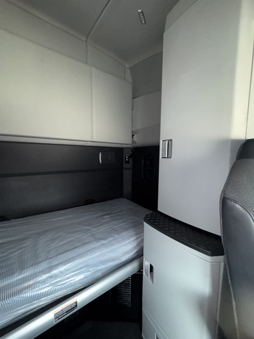 Kenworth T680 sleeper cab interior showing a bunk bed with striped mattress, overhead cabinets, and a vertical storage cabinet near the cab.