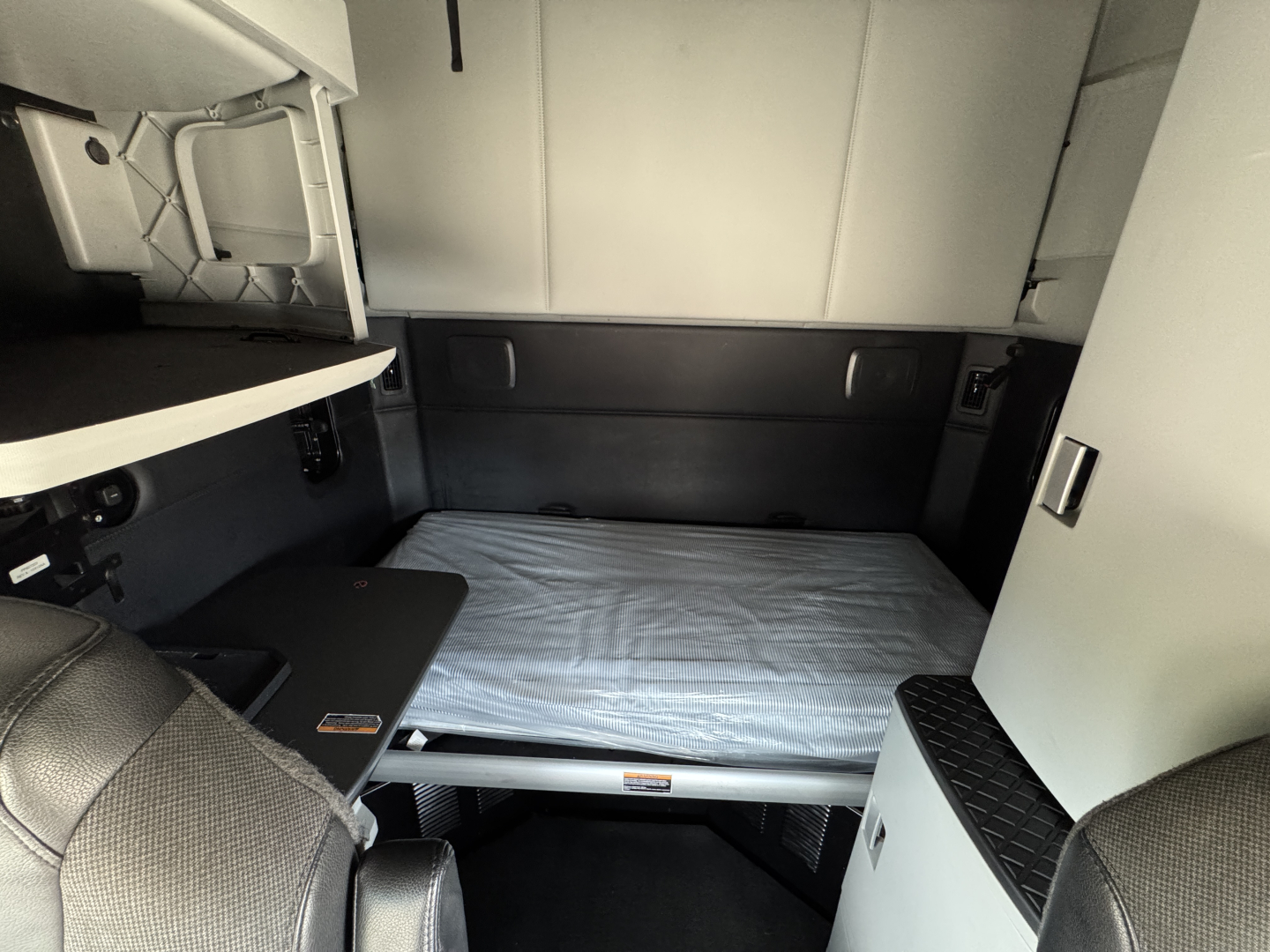 Kenworth T680 sleeper cab interior with a bed/cot folded into the wall, side storage cabinet, and compact work surface; light gray and black tones.