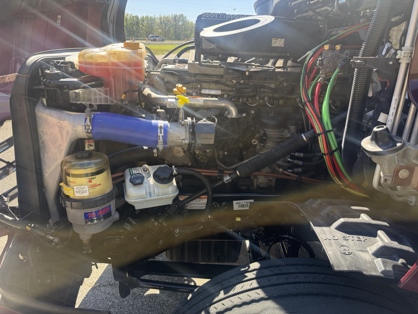Engine bay of a Freightliner Cascadia 126 truck showing coolant reservoir, blue hose, various fluid reservoirs, wiring harness, and turbo area.