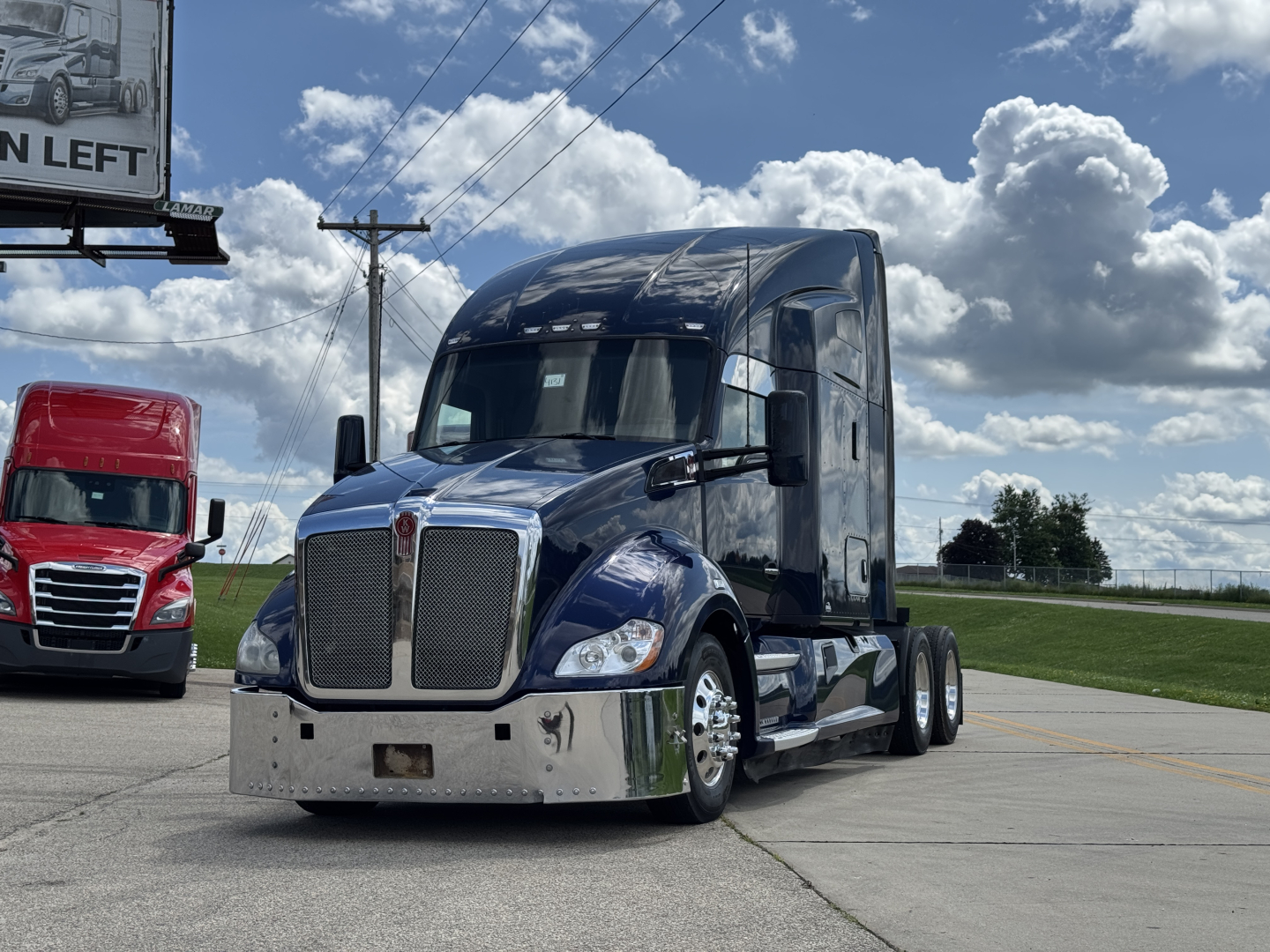 Kenworth T680 conventional sleeper truck, blue and chrome front, parked on lot with other truck in background.