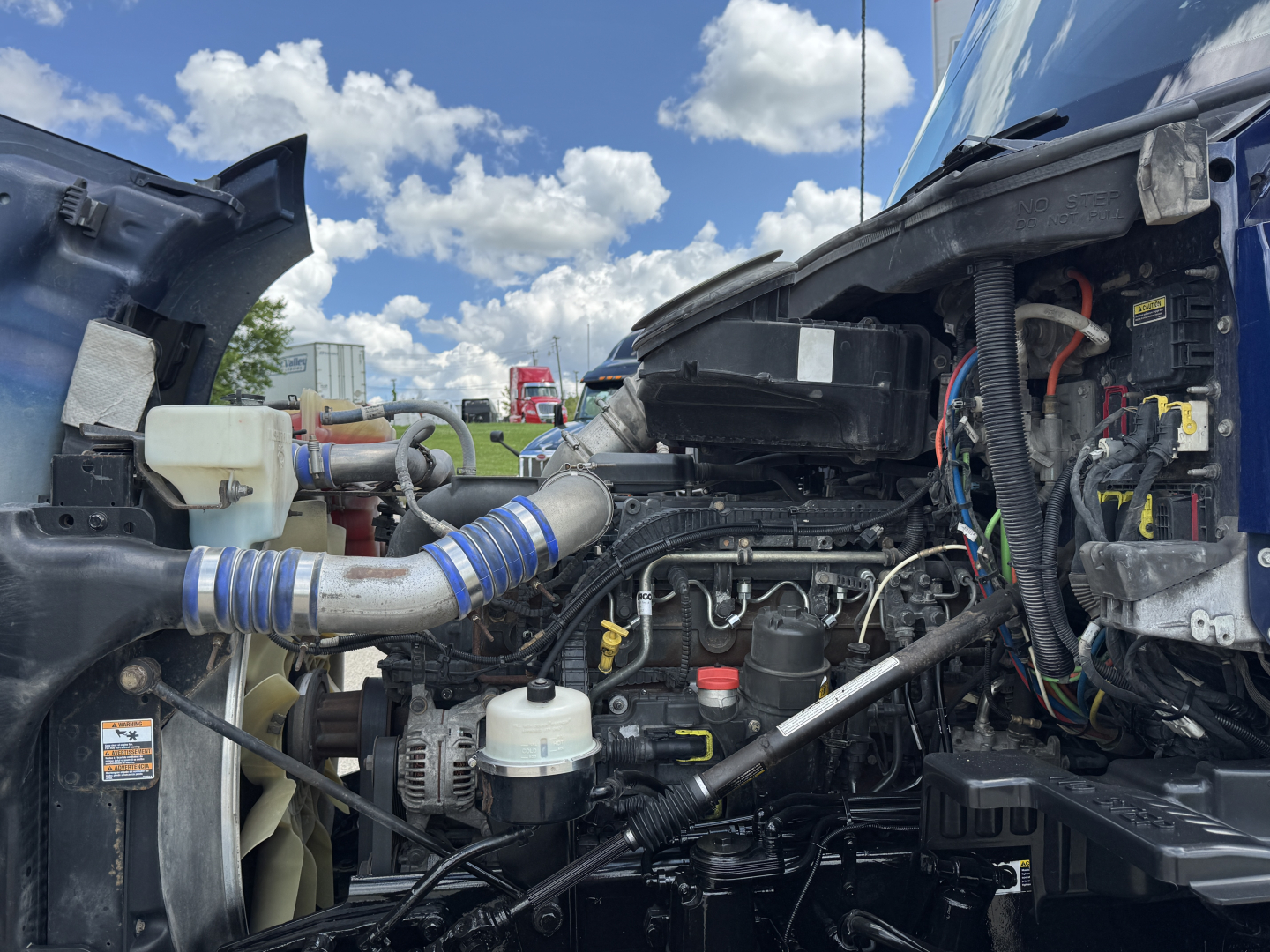 Kenworth T680 engine bay with visible hoses, wiring, and components under hood; coolant reservoir and intake hoses visible.