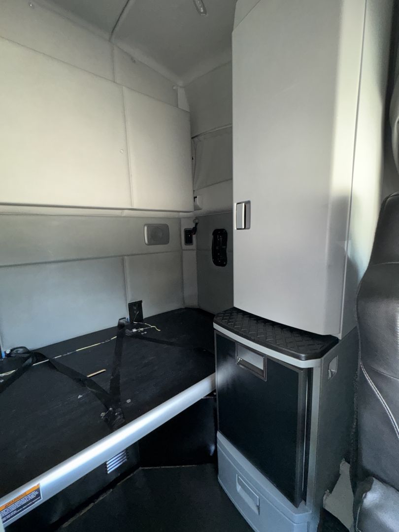 Kenworth T680 cab interior showing sleeper area with storage cabinet, small refrigerator, and seating; side wall panels and a work surface visible.