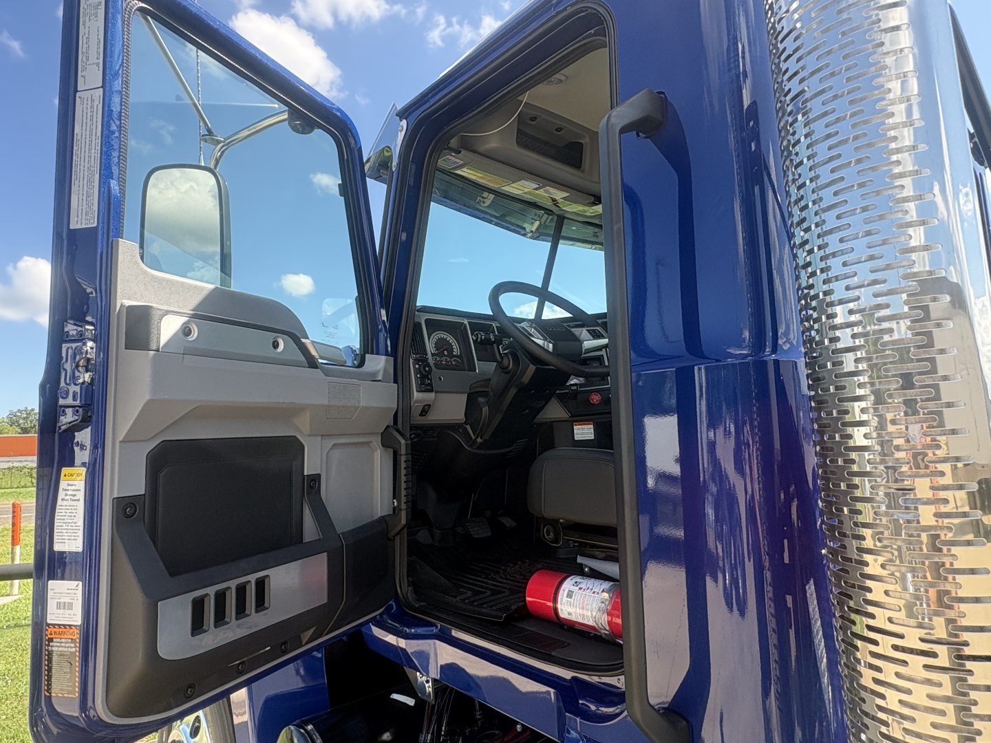 Mack PI64T cab interior and open driver door showing dashboard and steering wheel, chrome diamond-plate exhaust area on the cab side, blue exterior paint.