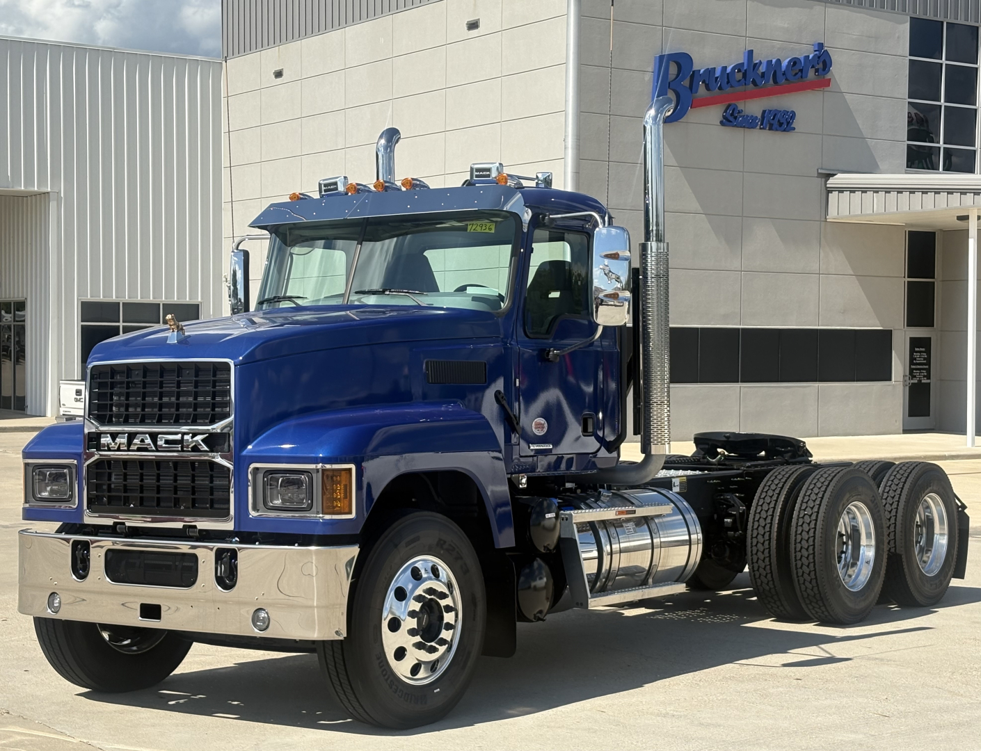 Mack PI64T conventional daycab truck in blue, showing front grille with MACK letters and chrome bumper, 6x4 chassis, at dealership.