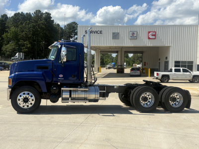 Truck chassis with blue cab and long chassis for a Mack PI64T. Visible are the blue cab, dual rear axles, silver fuel tanks, and black chassis with various components. In the background is a service center with signs and a few parked cars.