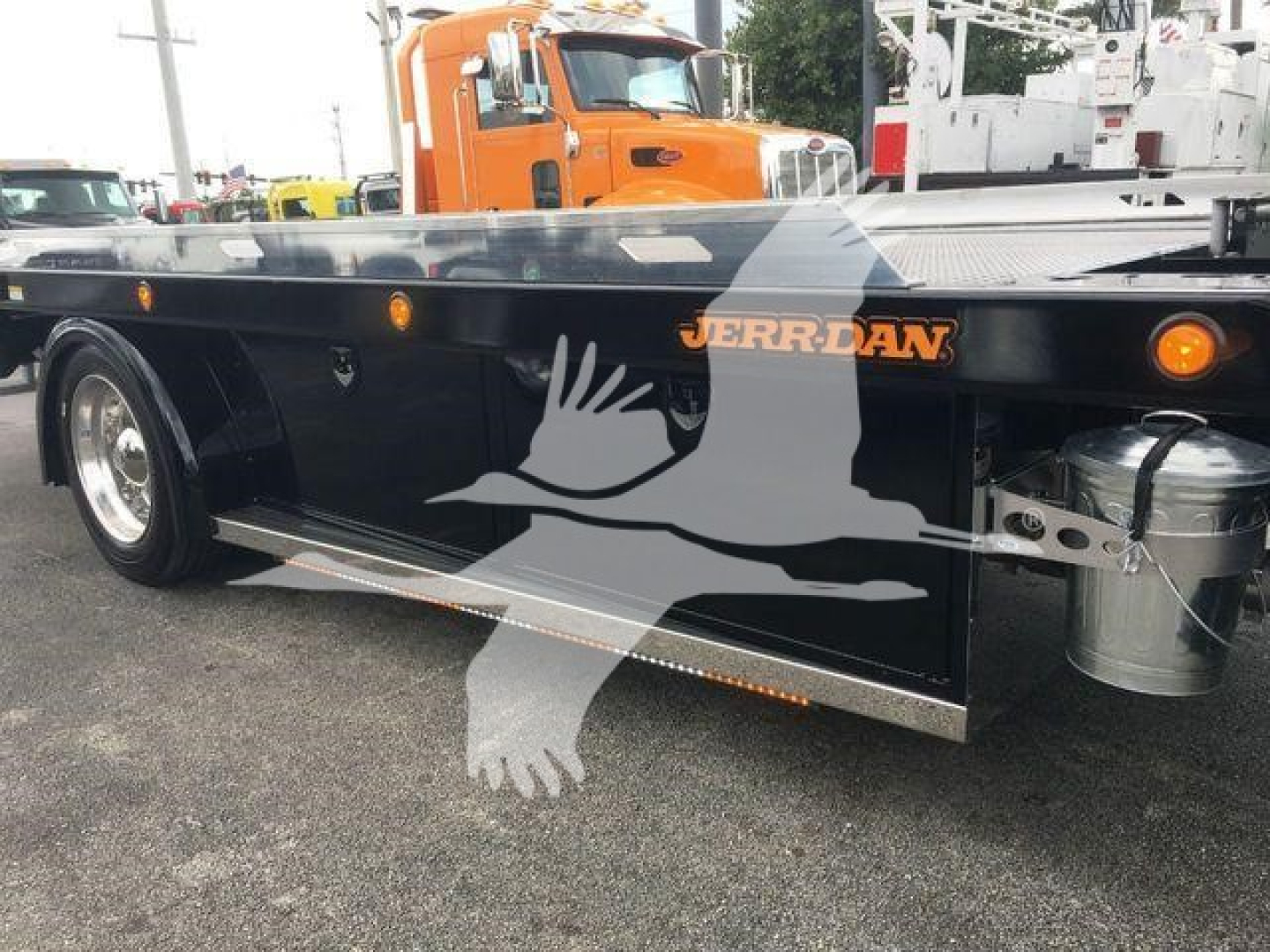 Peterbilt 337 rollback side view showing Jerr-Dan logo on the flatbed and a crane hook box; wheel, black bed, and orange cab visible.