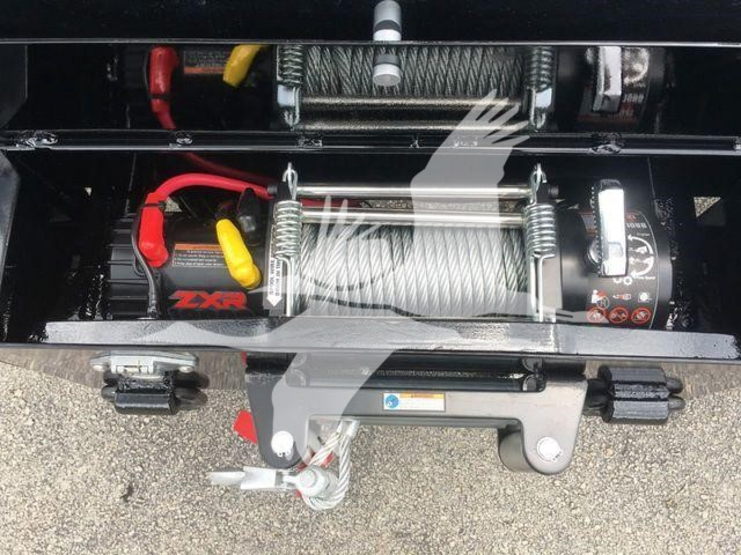 Truck bed winch assembly with steel cable, hook, and motor visible; power leads red and yellow; label stickers on winch; handle and mounting brackets in foreground.