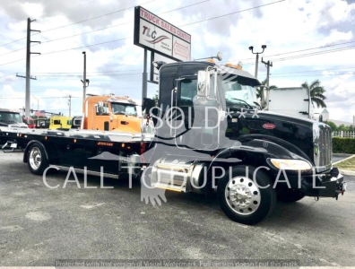 Peterbilt 337 truck in showroom/lot, black cab with rollback flatbed trailer attached, fuel tanks visible, signage in background, watermarked SOLD text.