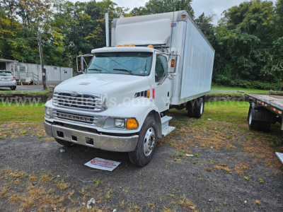 Box truck cab in front view with white Sterling Acterra nameplate, large chrome grille, and box trailer attached. Road grime on chassis, unused parking lot.