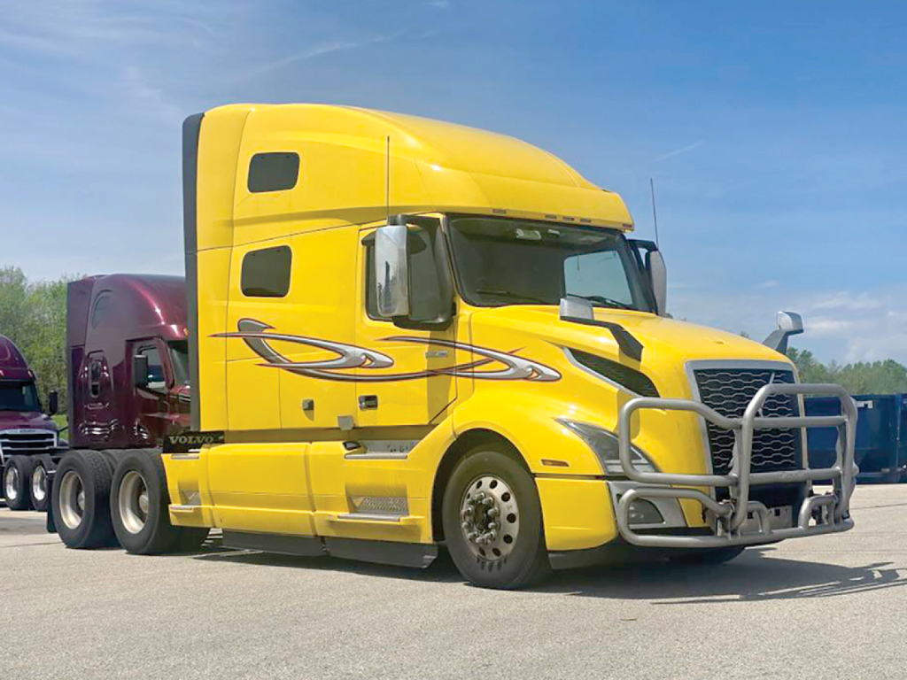 Volvo VNL 760 sleeper cab truck in yellow, front grille guard, side emblem graphics, on a trailer in a truck yard.