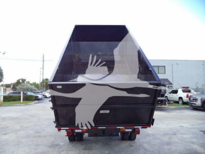 Truck bed with dump/roll-off container mounted on a Ford F550 chassis; rear view showing black enclosure with white graphic, trailers and vehicles in background.