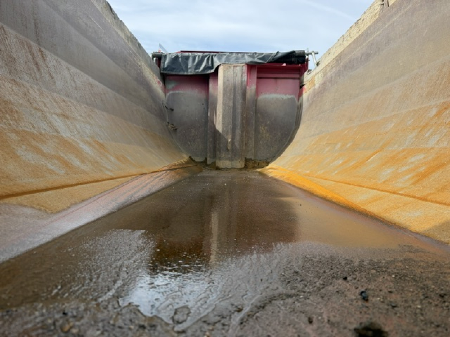 Truck dump bed interior showing wet, reflective metal sides with a red mechanism at the far end.