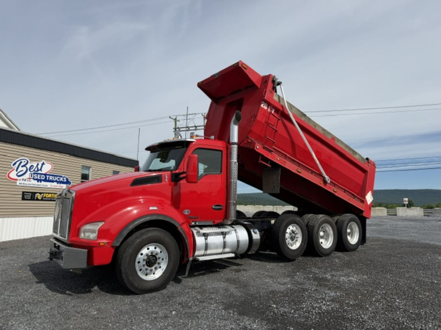 Red Kenworth T880 dump truck with raised dump bed, three axles, chrome fuel tanks, and black chassis. Visible text includes Best Used Trucks sign in background.
