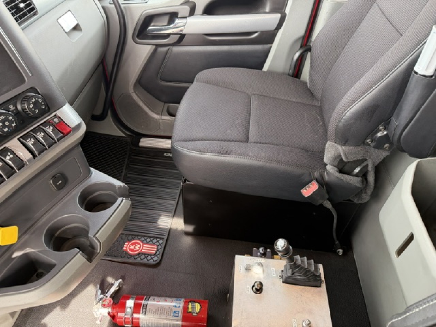 Kenworth T880 cab interior showing driver seat with center console and gear shift, floor mats, and a red tool/valve near the seat. 