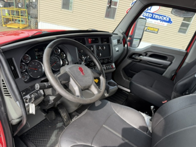 Cab interior of a red Kenworth truck showing steering wheel, dashboard with gauges, gear shifter, and front seats; visible window and outside sign indicate used trucks lot.