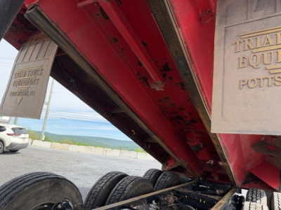 Kenworth T880 dump truck undercarriage showing red frame and dual rear axles with tires and a metal plate with embossed text.