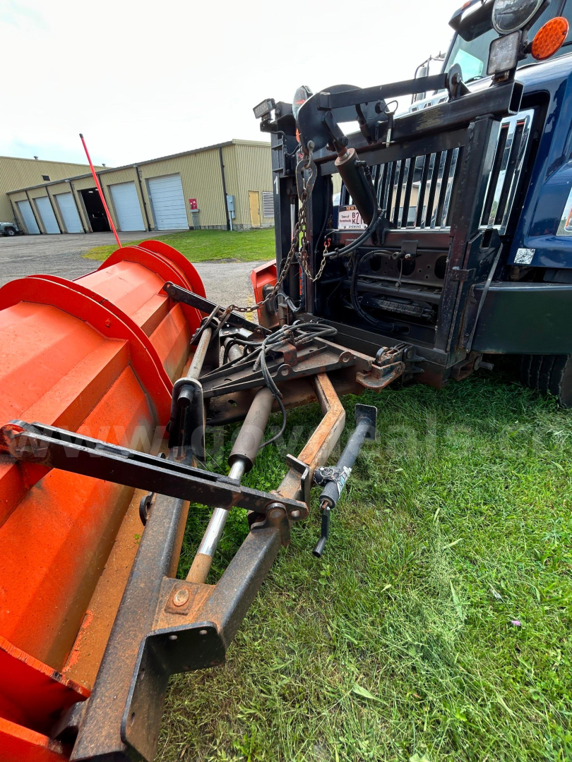 Equipment close-up showing a plow attachment connected to a blue truck’s front, hydraulic cylinders, and linkages.