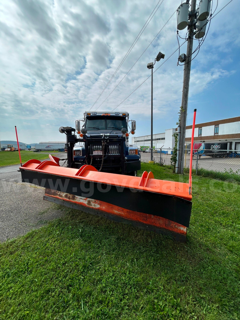 Front view of a black Western Star truck with a large orange snow plow blade attached, plow arms and red end plates visible, utility pole and highway behind.