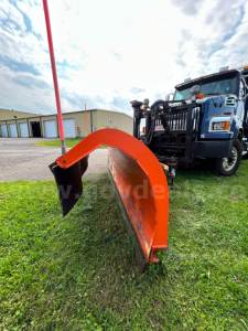 Front view of a 2018 Western Star 4700 with an orange snow plow attachment in the foreground, blue cab, and clear sky.