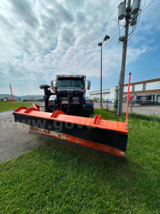 Front view of a black Western Star truck with a large orange snow plow blade attached, plow arms and red end plates visible, utility pole and highway behind.
