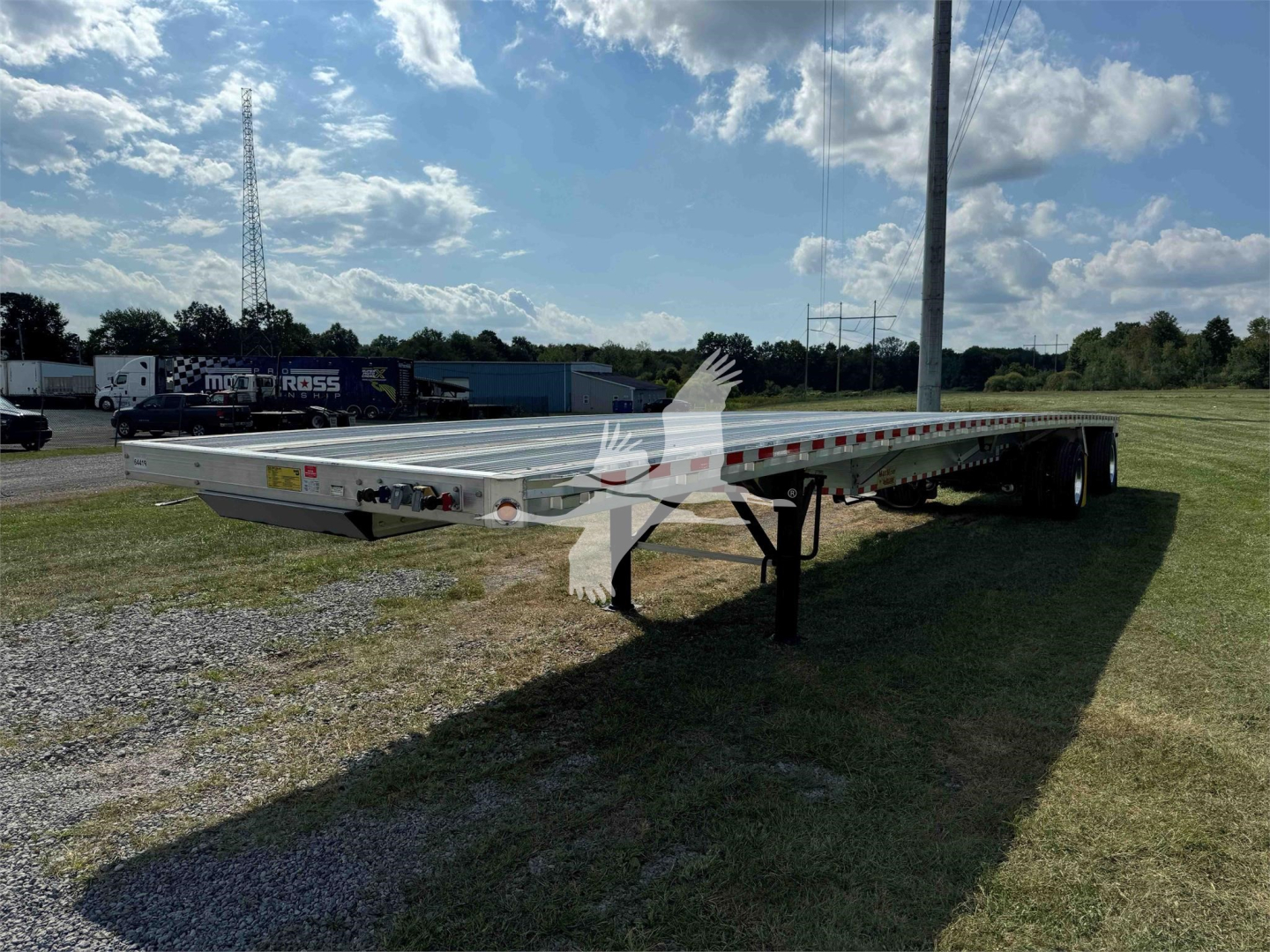 Flatbed trailer in side view showing dual axles and extended deck with reflective tape and support legs; sky and grassy field background.